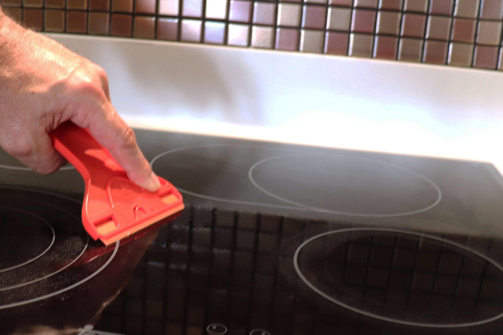 Hand using red Scraperite plastic scraper (SRTWGPO) on glass stove top for safe cleaning